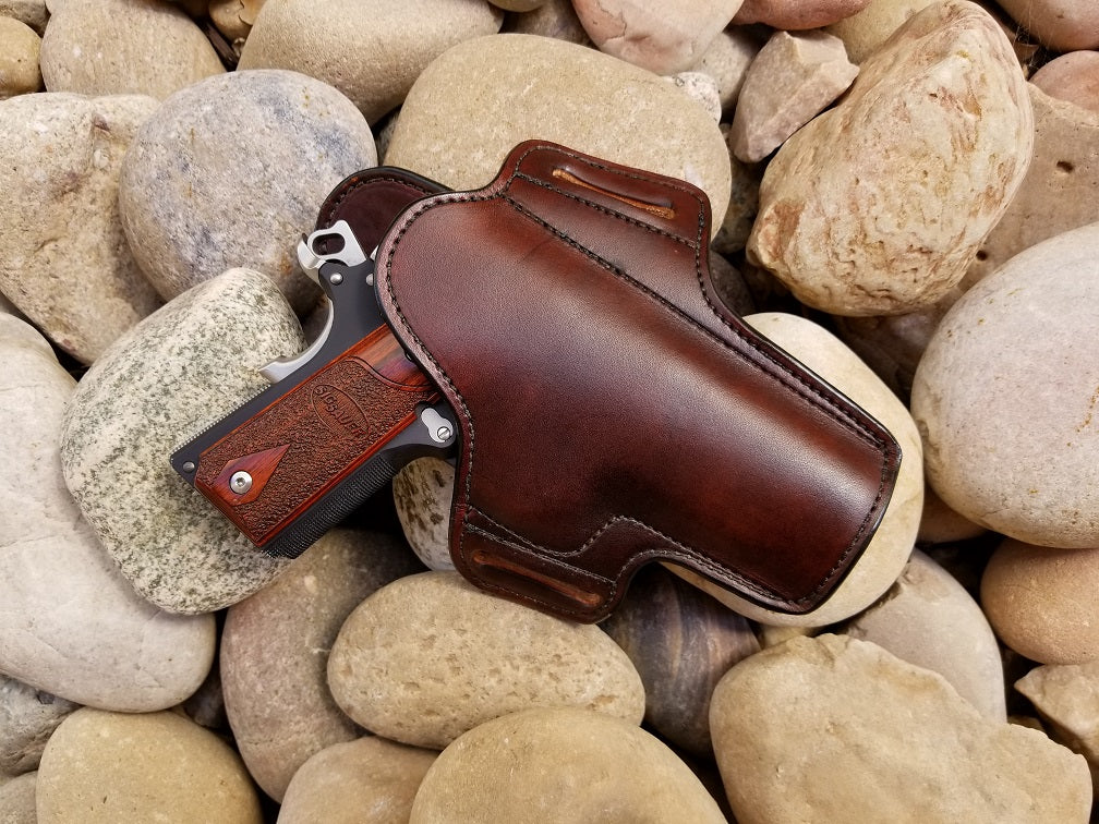 Rich brown leather pancake style holster for 1911 pistols sits on a bed of river rocks. 