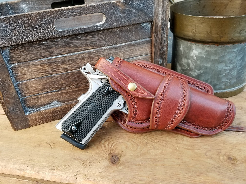 1911 western leather holster, in brown leather and tooled along the edges in a classic western style, sitting  next to a wooden crate and aluminum tin.