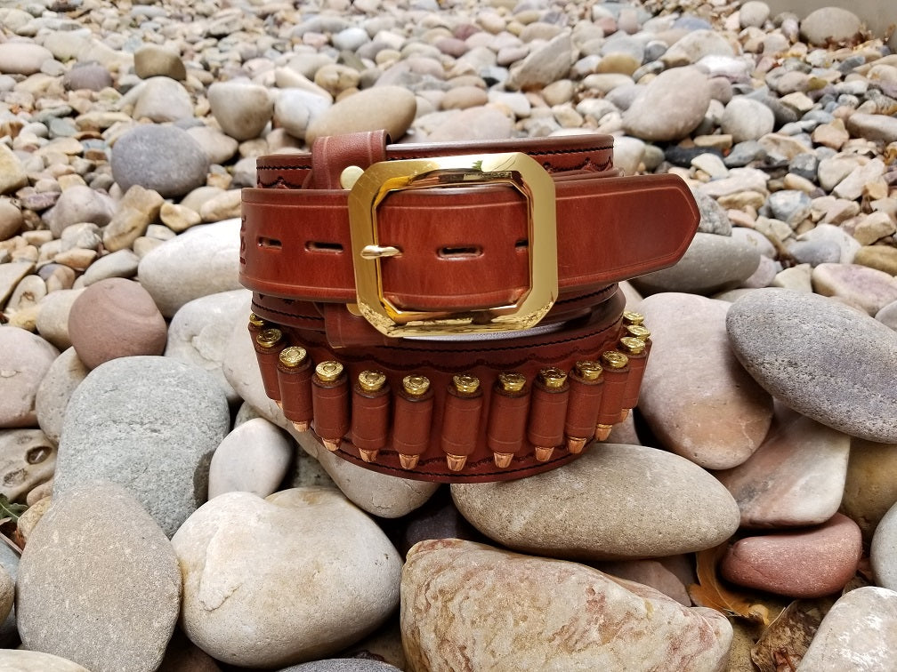 Brown leather cowboy gun belt with a bright brass buckle coiled on smooth gray river rocks