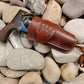 Beautiful smooth brown leather single action holster with a laced collar and medium drop sitting on smooth river rocks. At the bottom of the holster is a bundle of leather lace for a leg tie.