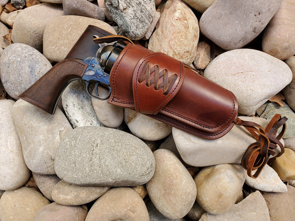 Beautiful smooth brown leather single action holster with a laced collar and medium drop sitting on smooth river rocks. At the bottom of the holster is a bundle of leather lace for a leg tie.