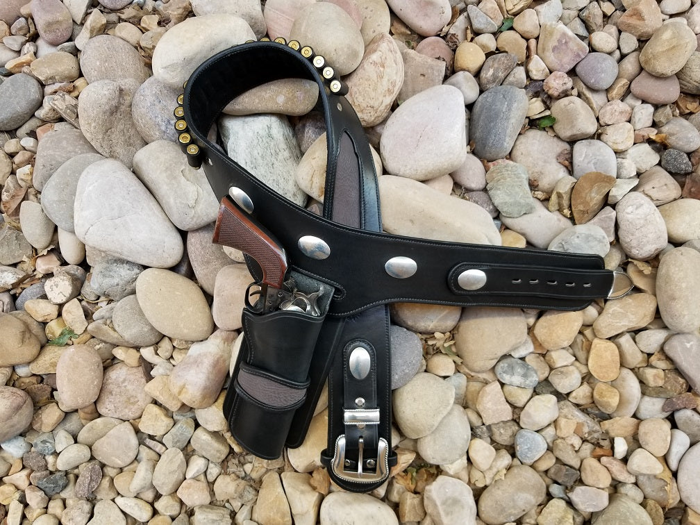 Stunning black leather buscadero drop rig sits halfway coiled on some river rocks. The black leather rig features large silver oval conchos and matt gray sharkskin inlay at the hips and across the front of the holster.