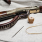 Lever action rifle sitting on white cloth next to a sinew spool and needle. Attached to the lever is a leather wrap and trigger guard made out of brown leather.