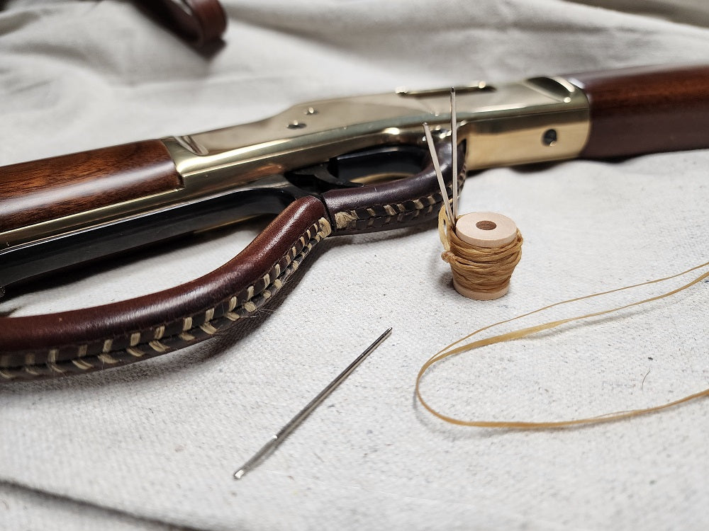 Lever action rifle sitting on white cloth next to a sinew spool and needle. Attached to the lever is a leather wrap and trigger guard made out of brown leather.
