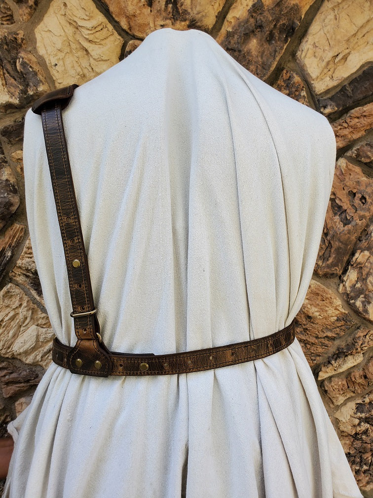 Back view of a brown distressed shoulder holster harness with waist and back strap visible. It features solid brass hardware and is fitted on a mannequin with jagged stone wall in background.