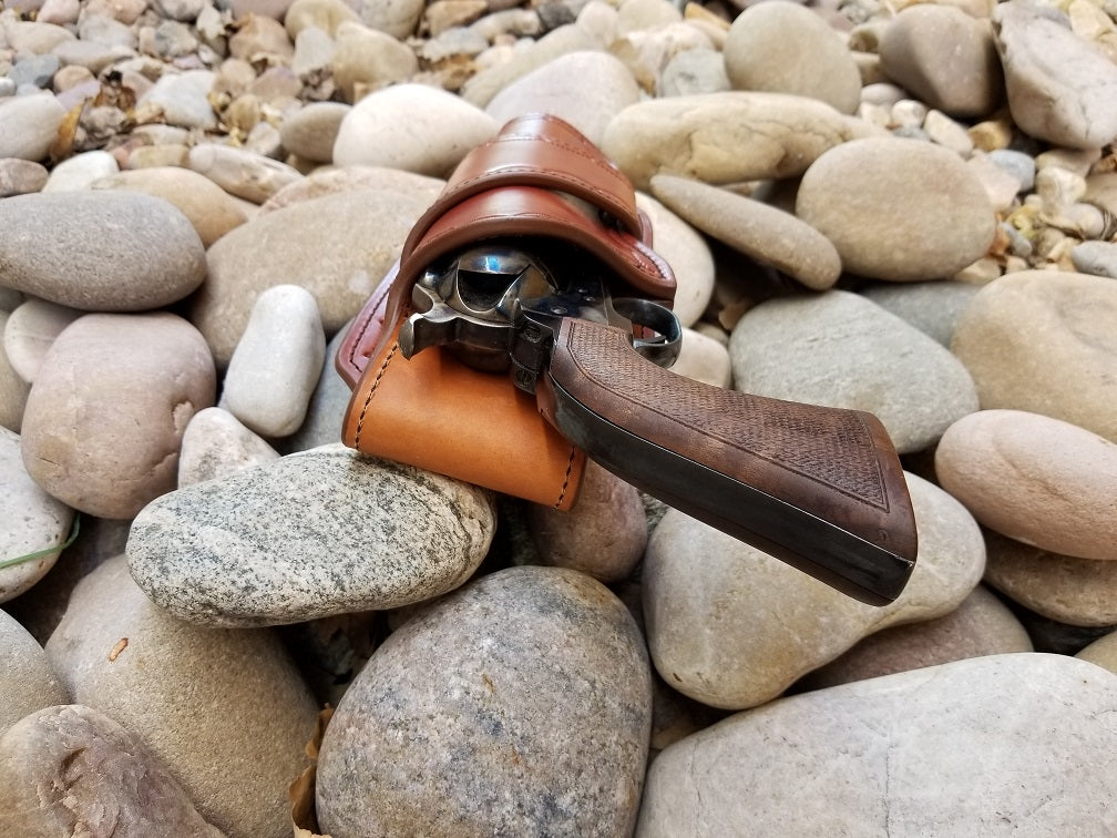 Rear view of the widow rock holster. The piece is a brown leather quickdraw style holster sitting on a bed of pale rocks. Holstered within is a Colt revolver.