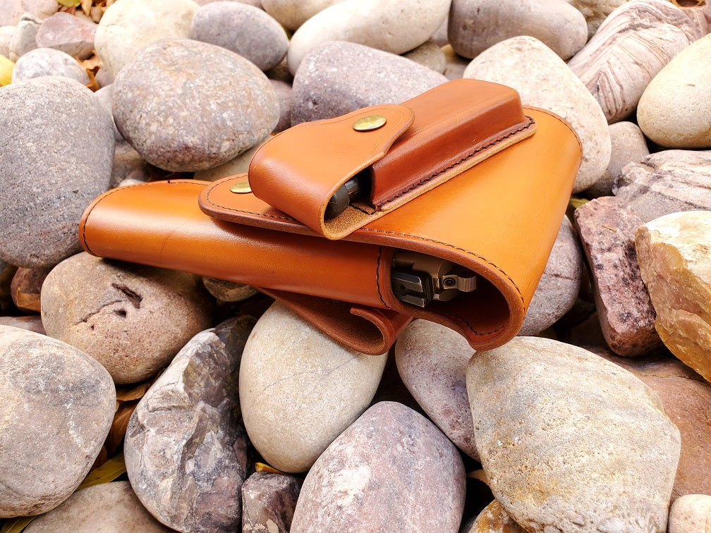 Top view of the russet leather vintage 1911 holster with flap coverage, on the flap is a magazine pouch. The holster is finished with solid brass hardware and black stitching, the piece lays on a bed of pale river rocks.
