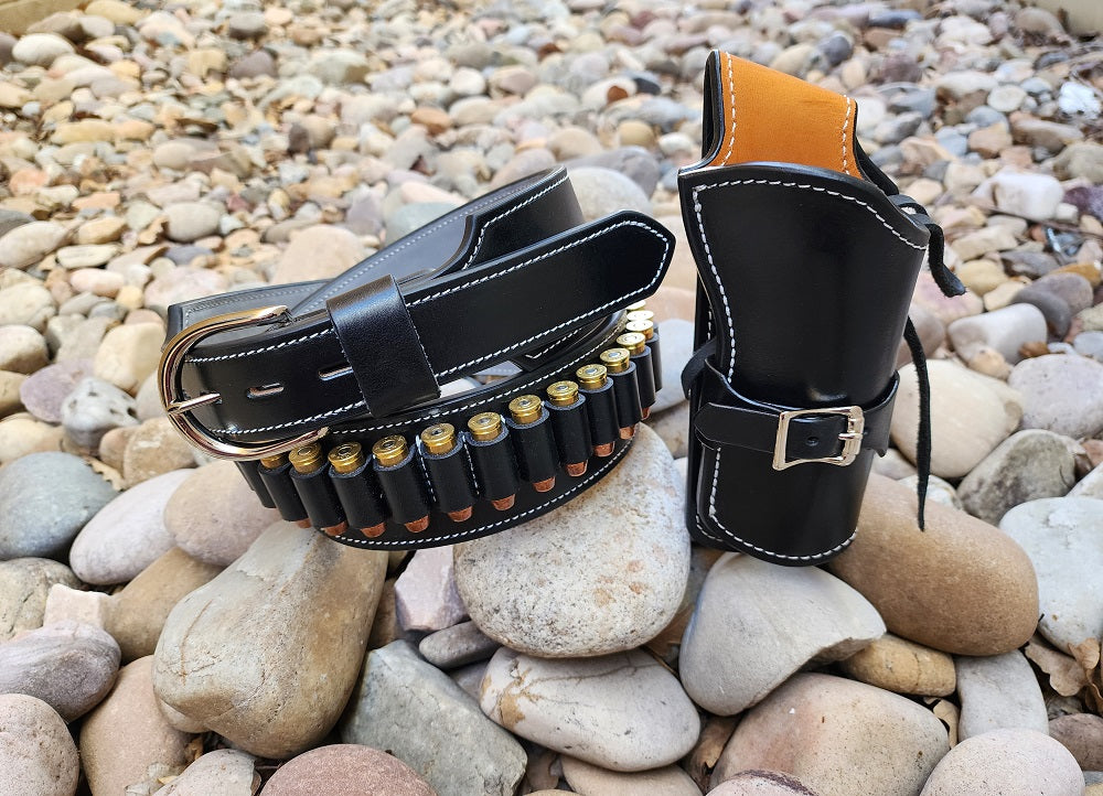 Black leather Angel Eyes inspired belt and holster, the belt has suede leather interior and 24 bullet loops and the holster has a russet leather interior. Both pieces are stitched with white thread and finished with solid brass silver hardware. Holster and belt and not attached in the image, they are both sitting on a bed of pale river rocks, the holster is standing up and the belt is curled up next to the holster.