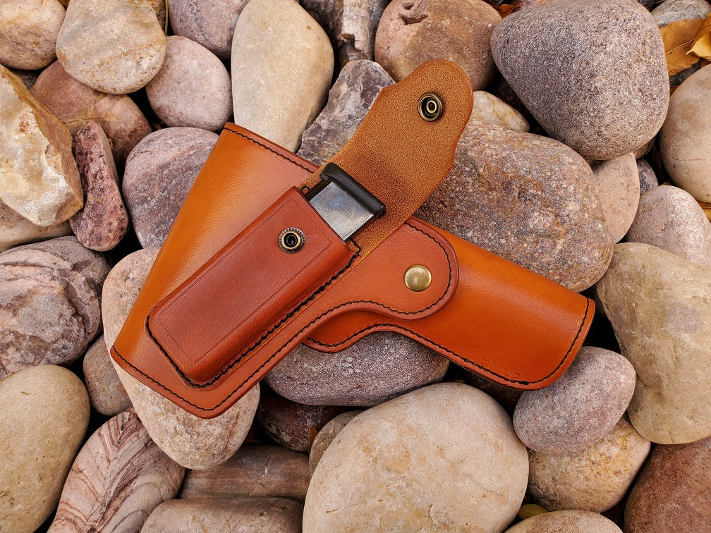 Russet leather vintage 1911 holster with flap coverage, on the flap is a open magazine pouch with a magazine inside. The holster is finished with solid brass hardware and black stitching, the piece is laying on a bed of pale river rocks.