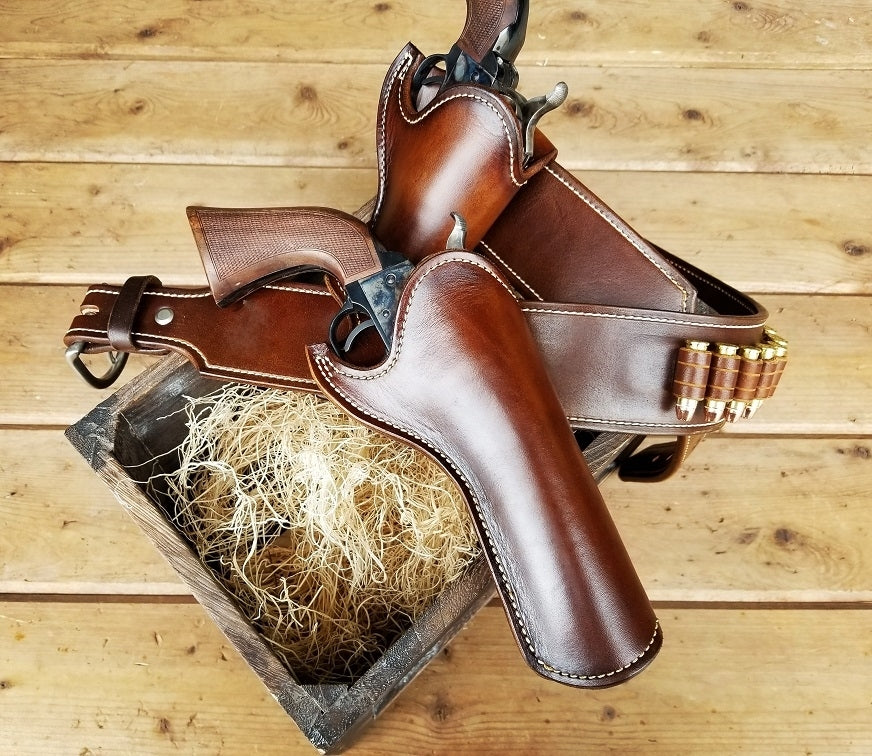 Brown leather gun rig with white stitching and 18 bullet loops with two holsters, the piece is finished with with solid brass nickel hardware. The gun rig lays on lightly colored wooden slats.