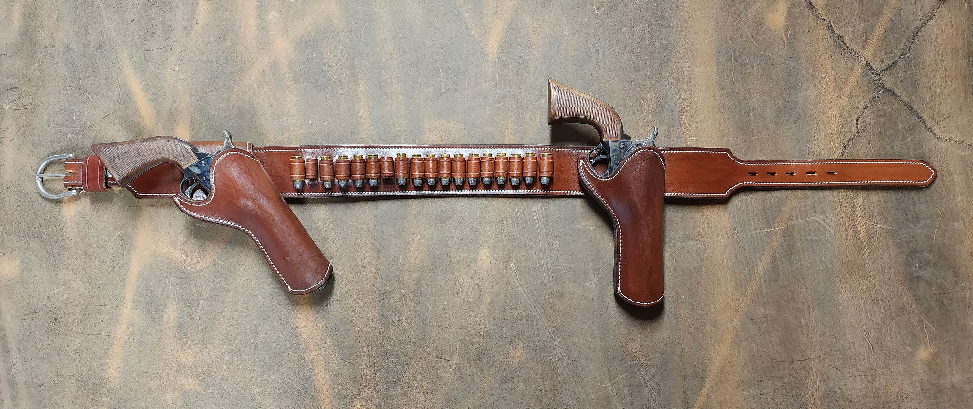 Josh Faraday inspired gun rig with right hand strong arm draw and right handed cross draw holsters attached to a brown leather belt with eighteen bullet loops. The piece is finished with white stitching and solid brass nickel hardware. The piece sits on a bed of ghostwood leather.