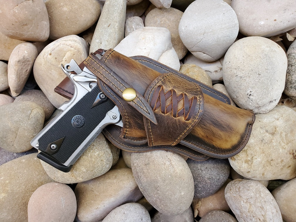 Handcrafted western holster for 1911 in rustic distressed brown leather and criss cross lacing sitting on a bed of pale river rocks.