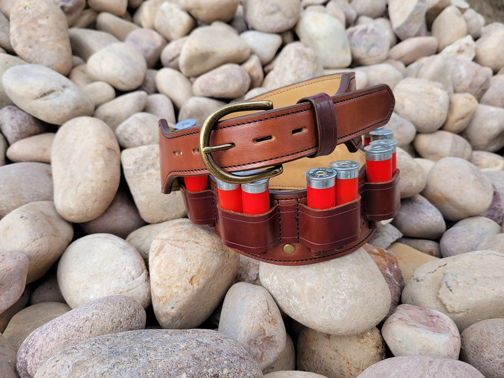Brown leather shotgun ammo belt filled with 12 gauge rounds in double pockets sitting coiled on smooth river rocks.