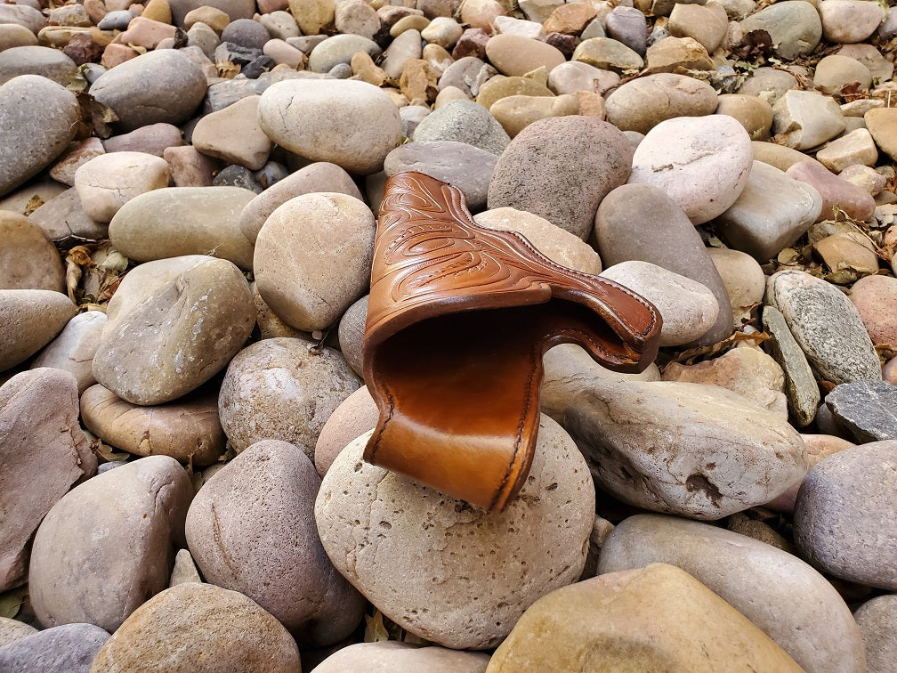 view from mouth end of a tooled brown leather holster sitting on a bed of pale river rocks. The holster features black stitching and intricate leaf pattern tooling.