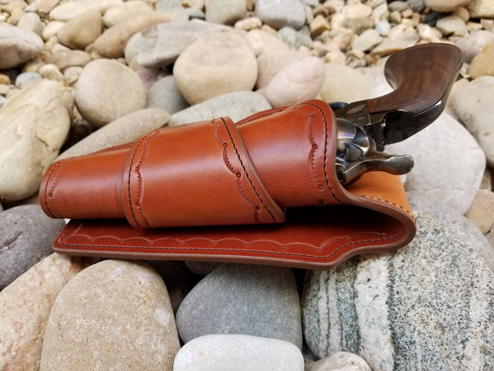 The is a brown leather holster and is tooled in a scallop pattern and sits on a bed of pale river rocks.